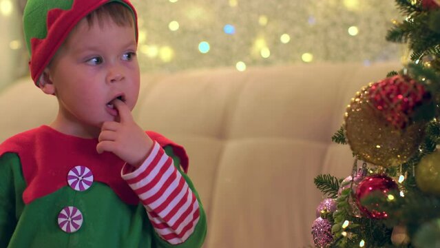 little elf boy put finger in mouth looks pensively at decorated Christmas tree admiring beautiful decorations and dreams Christmas and new year celebrations. child in green elf costume Santa's helper