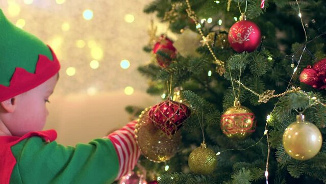 little child boy in elf costume decorates Christmas tree with beautiful decorations preparing to celebrate Christmas and New Year in room. kid Santa helper hanging baubles on Christmas tree branch