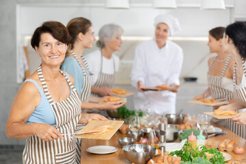 Mature woman holding cutting board with raw chicken breast in her hands posing surrounded by other members of cooking course