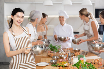 Young girl holding bowl and whisk in her hands surrounded by other female members during cooking master class