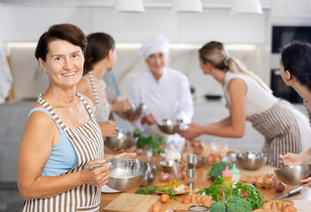 Mature woman at group cooking class holds bowl in hands and mixture cream sauce with white wine for meat with whisk