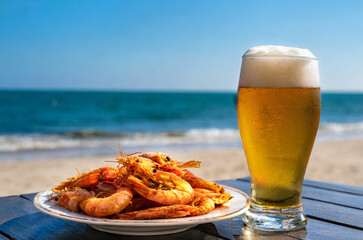 A glass of beer and a portion of fried prawns on a table at the beach - obrazy, fototapety, plakaty