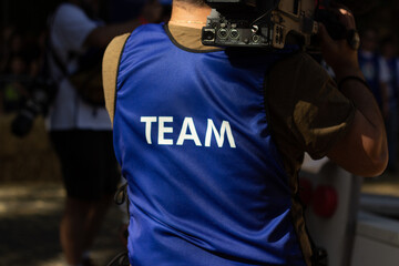 Fototapeta premium Tbilisi, Georgia - October 22, 2023. Back view. Close up videographer in blue vest with text Team with camera. Concept of fun, holiday, joy, tv operator, video, cinema, movie maker, film, shoot