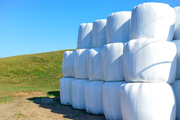 Rows of white plastic covered round bales of hay stacked three high on a farmer's field. The silo...