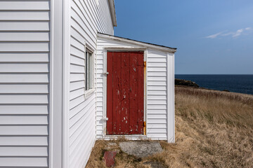The corner of a white wooden traditional house with a small double hung window and a wooden vibrant red colored screen door. There's yellow-colored grass and a blue ocean in the background. 