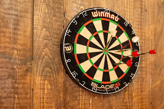 St. John's, Newfoundland, Canada- October 2023: Regulated Winmau dartboard hanging on a brown wooden wall.  There are three red darts and one white dart on the board with wire numbers and a bullseye. 