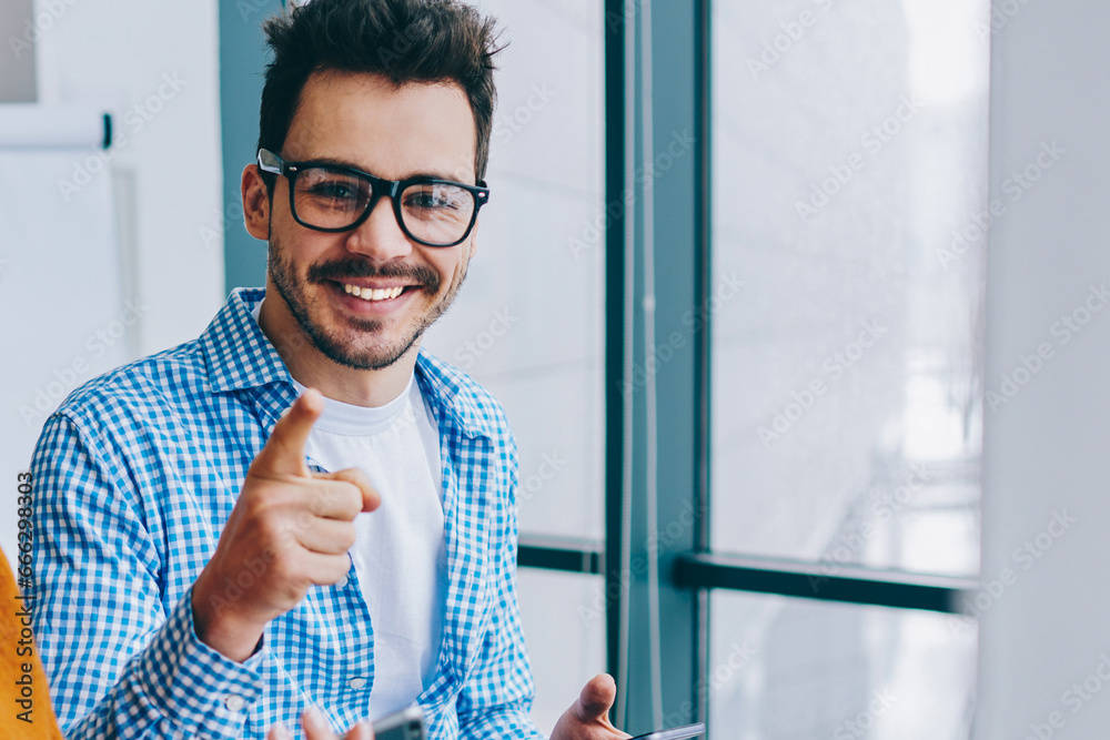 Wall mural portrait of cheerful millennial hipster guy in optical spectacles pointing while smiling at camera