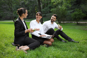 Happy colleagues with laptop having business lunch on green grass in park