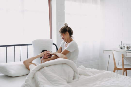 Funny asian couple wife try to wake lazy husband up in white bedroom.