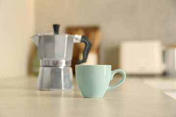 Cup of coffee and moka pot on light table in kitchen, selective focus