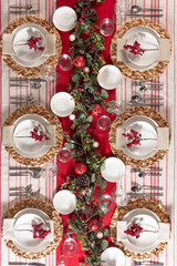 Festive Christmas dining table setup with elegant white plates adorned with bright red berry branches, set on woven placemats, amidst a vibrant red table runner, on a striped fabric backdrop, top view