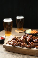 Tasty chicken wings, ketchup, glasses of beer and vegetable sticks on wooden table. Delicious snacks