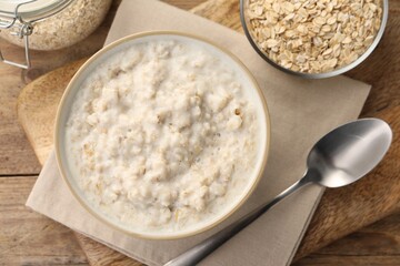 Tasty boiled oatmeal in bowl, flakes and spoon on wooden table, flat lay