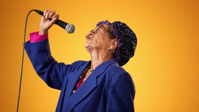 Profile of funny crazy toothless grandmother with a fashionable look with glasses, singing enthusiastically into a microphone and dancing isolated on solid yellow background