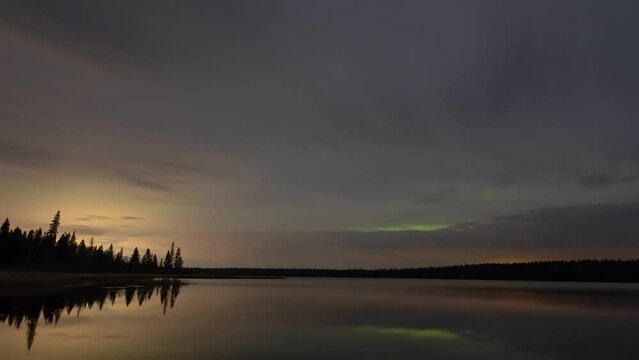 Gray nighttime clouds with the occasional colorful patch of Aurora move towards the viewer above a calm lake in theis time lapse. The scene is near perfectly reflected in the water.
