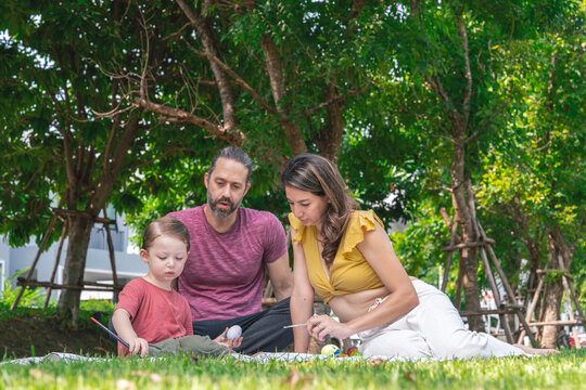 family has a picnic in the garden. Parents and their three-year-old son sit and play together. Father and mother do activities to teach their son to paint together in the garden