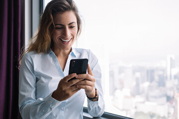 Adult woman texting on phone nearby window smiling