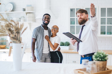Confident estate agent showing kitchen to African American pleased hugging couple - Powered by Adobe