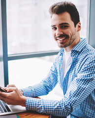 Portrait of young cheerful skilled male copywriter looking at camera and using smartphone