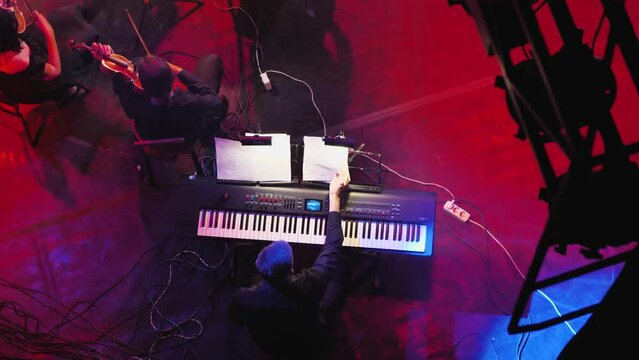 A man at a synthesiser shifting notes while violinists play nearby. Keyboardist in the symphony orchestra. View from above. Light accompaniment of the show