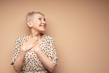 A cheerful elderly woman in a polka dot dress rejoices, pressing her hands to her chest. Delight...