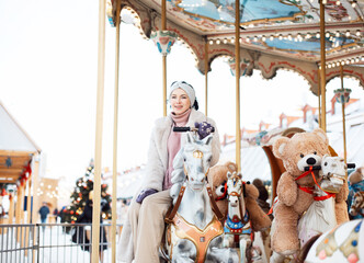 Cheerful woman in a faux fur coat and mittens rides a carousel and has fun