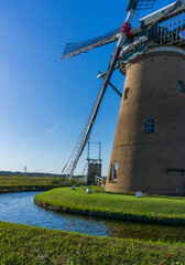 Fototapeta premium A picturesque windmill stands tall against a clear blue sky, its sails casting shadows. Surrounded by pristine waters and lush greenery, it embodies the tranquil beauty of pastoral scenes. 