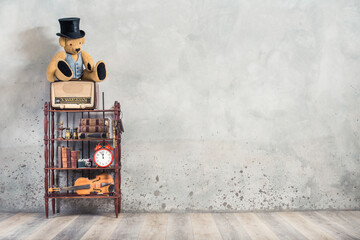 Classic radio, vintage Teddy Bear in cylinder hat, antique books, alarm clock, camera, binoculars, fiddle, keys on shelf front concrete wall background. Retro old style filtered photo