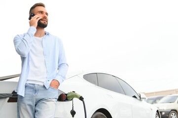 Beautiful young stylish man is with electric car at daytime charging the vehicle