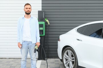 Beautiful young stylish man is with electric car at daytime charging the vehicle