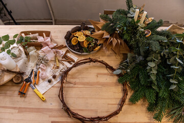 Master class, instructions, DIY how to make Christmas wreath for Advent. On wooden table: fir branches, eucalyptus, wreath base, ribbons, scissors, dried oranges. Eco-friendly home decoration