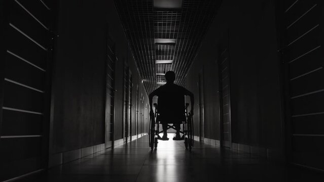 Tilting down monochromatic shot of female wheelchair user going through long dark hallway in clinic