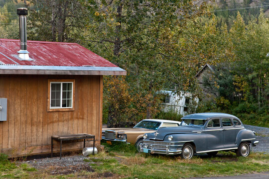 Two classic vintage cars parked beside a wooden house in Chitina, Alaska