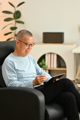 Smiling senior man in glasses using digital tablet while sitting on armchair at home