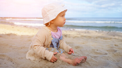 Cute little girl sits on clean sand on beach. Toddler next to clear sea playfully burying feet in warm sand enjoying perfect day by shore, sunlight