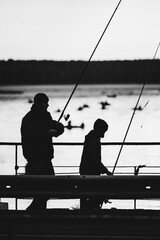 Silhouette of a teenager with his father on a bridge with fishing rods against the background of a water surface with boats with fishermen..