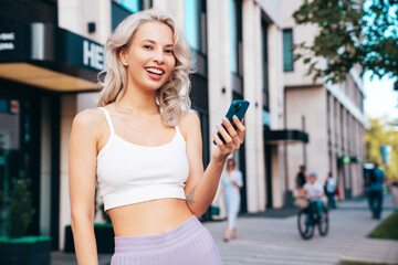 Young beautiful smiling hipster woman in trendy summer clothes. Carefree model posing in street at sunset. Positive blond female. Holds smartphone, uses phone apps, looks at telephone screen