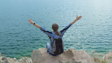 Female tourist with backpack on her shoulders sits on rock above sea with her hands raised up.