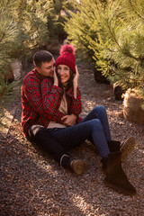 Close-up portraits of young couple in red checkered shirts sitting among Christmas tree seedlings at street market. Man embrace woman in knitted hat with love. Warm winter date.