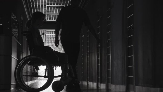 Full shot of unrecognizable male figure reassuring his wife on wheelchair in long dark hallway of hospital