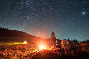 Night camping in mountains under starry sky. Man sitting on grass near fire, holding stick. Male...
