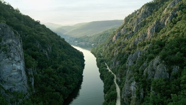 Scenic view of the Danube River, flanked by lush green mountains with the iconic rock sculpture of Decebalus in Dubova, Romania.