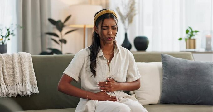 Woman, upset and stomach pain for digestion in home with discomfort on sofa in living room. Indian person, bad diet and nutrition with problem, trouble or stress for health, wellness or self care