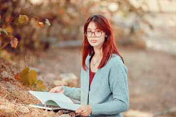 Red-haired student girl is studying outside, putting notebooks on the grass.