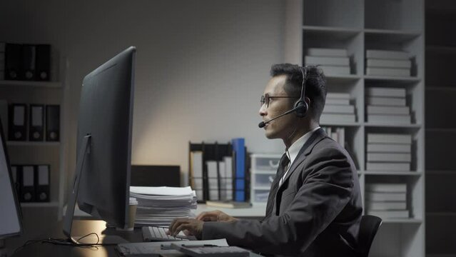 Businessman wearing headset sitting in office using desktop computer talking with customers in video conferencing app. Remote help, virtual meeting by business, negotiation concept