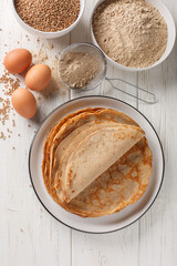 Traditional French Savory Buckwheat Galettes Bretonnes pancakes on a table with flour, cereal and eggs closeup on the white wooden table. Vertical top view from above - obrazy, fototapety, plakaty