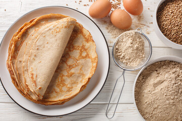Buckwheat crepe, galette on a plate with ingredients flour, cereal and eggs closeup on the white wooden table. Horizontal top view from above - obrazy, fototapety, plakaty