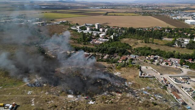 Aerial footage of a fire burning and spreading along a garbage field next to the largest residential neighbourhood of Bulgaria. Lyulin illegal dump on fire. Firefighters putting out fire. Drone shot.
