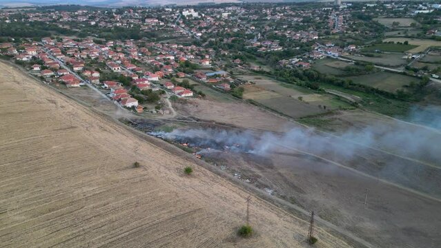 Aerial footage of a fire burning and spreading along a garbage field next to a small village. Illegal arson of a dump. Drone shot.
