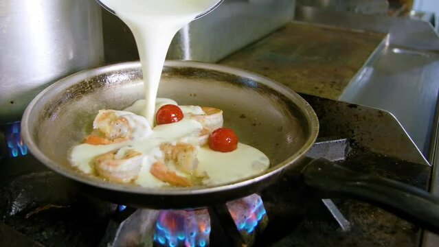 Chef drizzles heavy cream into pan on gas stove with shrimp , omatoes and milky white suace